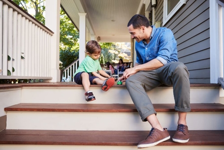 Father And Son Sit On Porch Of House Playing With Toys Together